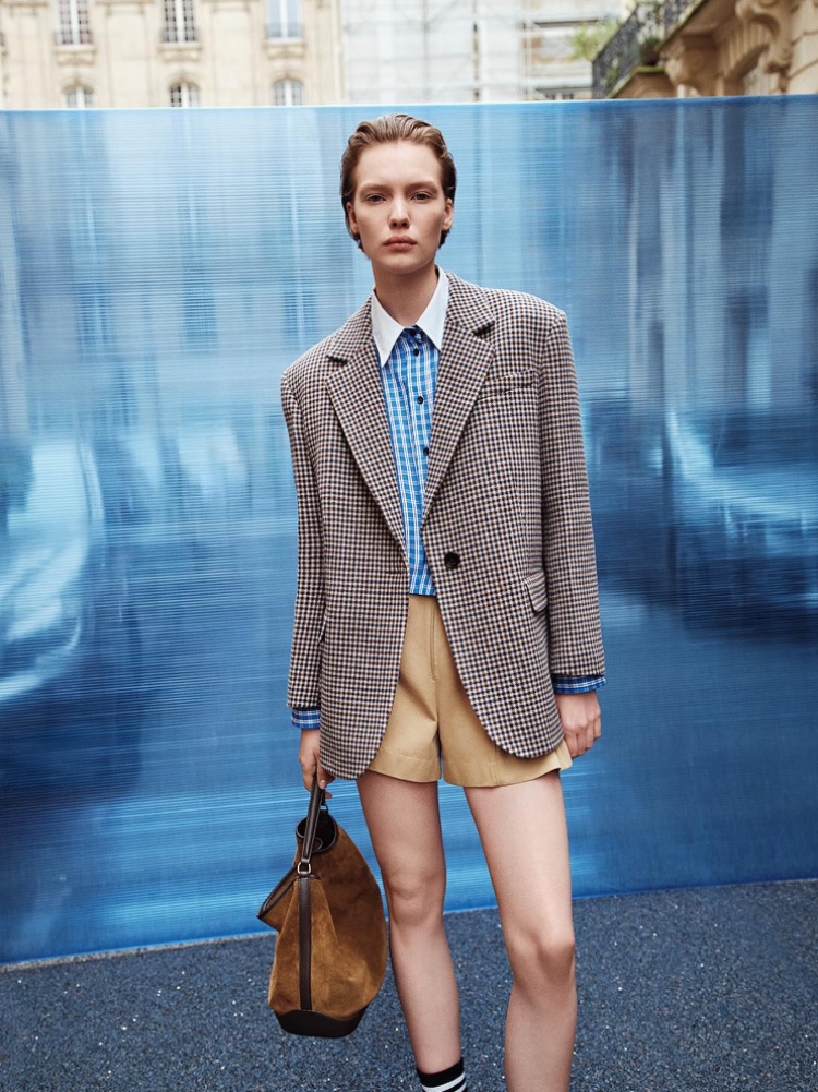 A model dressed in a checked shirt, jacket, and ruffled shorts, holding a suede bag from Sandro